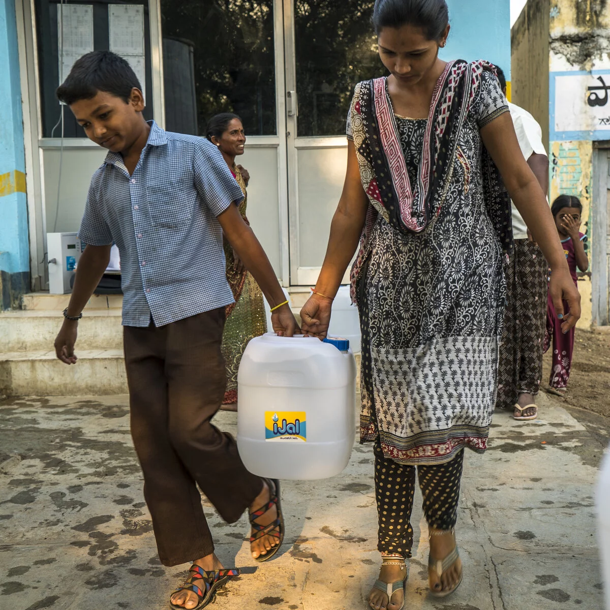 Neighbours help each other carry the 20 litre water cans home after buying safe water from the iJal station in Peddapur, a remote village in Warangal, Telangana, India, on 22nd March 2015. Safe Water Network works with local communities that live beyond the water pipeline to establish sustainable and reliable water treatment stations within their villages to provide potable and safe water to the communities at a nominal cost. Photo by Suzanne Lee/Panos Pictures for Safe Water Network 2025/09/20150322-SafeWater-Warangal-0250_1x1.jpg